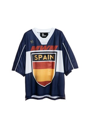 Joven con cabello trenzado lleva una camiseta deportiva de España de gran tamaño, pantalones cortos beige hasta la rodilla, calcetines blancos y botas marrones sobre fondo liso.