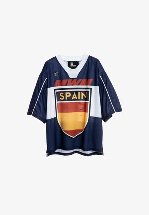 Joven con cabello trenzado lleva una camiseta deportiva de España de gran tamaño, pantalones cortos beige hasta la rodilla, calcetines blancos y botas marrones sobre fondo liso.