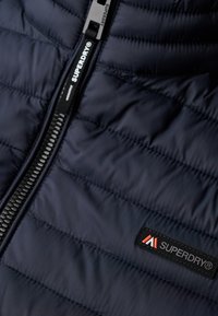 Granatowa pikowana kurtka z pełnym zamkiem błyskawicznym, wyposażona w czarną naszywkę z logo Superdry oraz szczegóły o teksturowanej powierzchni. Podszewka wydaje się gładka i elegancka.
