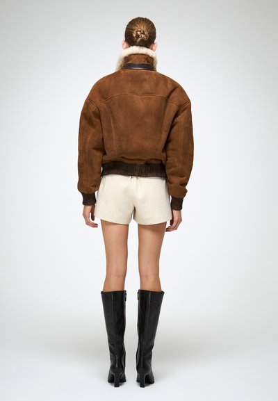 Giacca bomber in suede marrone con colletto peloso, polsini e orlo rigati; abbinata a pantaloni corti crema e stivali neri alti fino al ginocchio.
