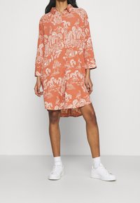 Terrakotowa sukienka koszulowa z białymi tropikalnymi wzorami, z przodem na guziki, kołnierzykiem i rękawami do połowy łokcia. Noszona z białymi trampkami.