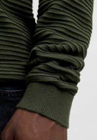 Pulover tricotat de culoare verde măslină, cu un model texturat de nervuri, având mâneci gathered și o manșetă strânsă, prezentat pe un fundal din stofă închisă.