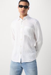 Mann mit schwarzen rechteckigen Sonnenbrillen, einem weißen Langarmhemd mit knöpfbarem Kragen und einer Fronttasche sowie einer hellblauen Jeans.