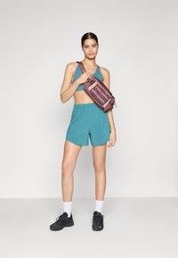 Top cropped atlético em teal e calções de corte solto. Saco de cintura roxo da Patagonia com logótipo, sapatos atléticos pretos, meias brancas até ao tornozelo.