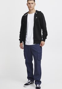Zwarte rits hoodie met zijzakken, witte T-shirt eronder, marineblauwe cargobroek met meerdere zakken en zwarte sneakers met witte accenten.