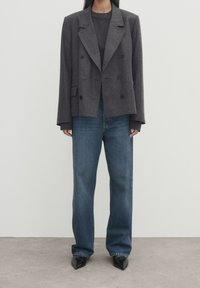 Blazer grigio a doppio petto su una camicia grigio scuro, abbinato a jeans blu larghi e scarpe nere a punta. Design minimalista, forma strutturata.