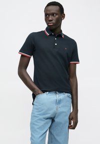 Ναυτικό πόλο μπλουζάκι με κόκκινο και λευκό ριγέ γιακά και μανίκια, διαθέτει τριπλό κουμπί και ένα μικρό κεντητό λογότυπο στο στήθος.