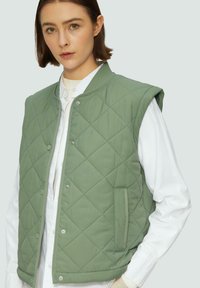 Gilet matelassé vert avec un col rond, fermeture à boutons pressions et poches latérales, superposé sur une chemise blanche. Détails de tissu texturé visibles.