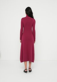 Φόρεμα με μακριά μανίκια και ψηλό γιακά σε βαθύ ματζέντα, εφαρμοστό στον κορμό, με φουσκωτή φούστα, ομαλή υφή, συνδυασμένο με μαύρα πέδιλα με τακούνι.