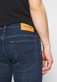 Dunkelblaue Jeans aus Denim mit einem Lederpatch mit der Aufschrift "DIESEL" auf der Rückseite. Verfügt über eingenähte Gesäßtaschen und kontrastierende Nähte.