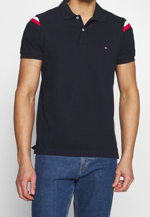Homme portant un polo noir à manches courtes avec des rayures rouges et blanches sur les épaules et un jean bleu, debout devant un fond uni.