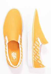 Des chaussures en toile jaunes à enfiler avec des semelles en caoutchouc blanches. Une des chaussures présente un motif en damier à l'intérieur du col et des détails de logo sur les côtés.