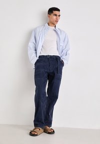 Lichtblauw gestreepte lange mouwen shirt, witte crew-neck t-shirt, marineblauwe cargobroek met zakken, en bruine sandalen met een teenlus.