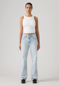 Λευκό αμάνικο κοντό τοπ και ανοιχτό μπλε τζιν παντελόνι με καμπάνα. Το τζιν έχει θηλιές για ζώνη και μπροστινές τσέπες. Το μοντέλο στέκεται μπροστά σε ουδέτερο φόντο.