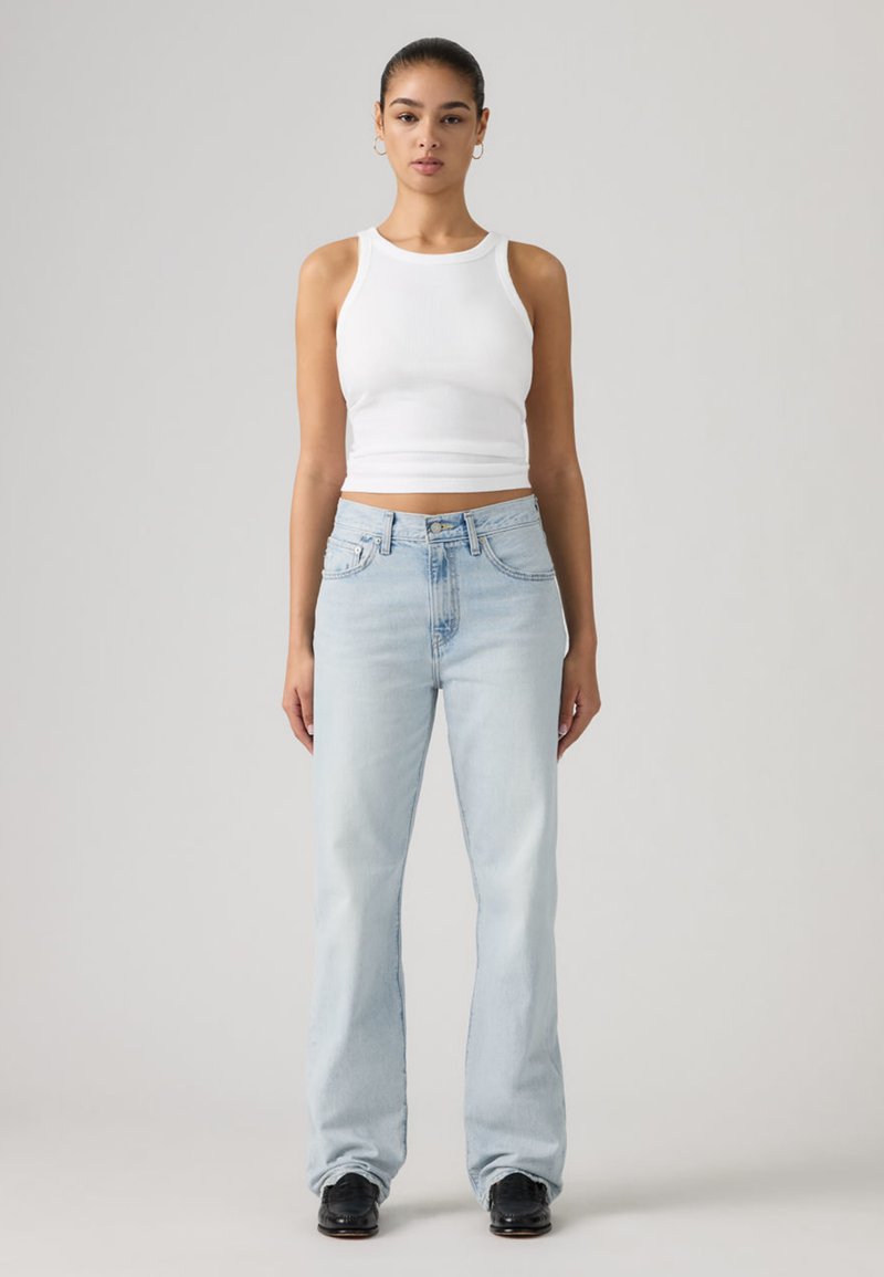 Haut court blanc sans manches et jean évasé bleu clair. Le jean comporte des passants pour ceinture et des poches avant. Le modèle se tient devant un fond neutre.