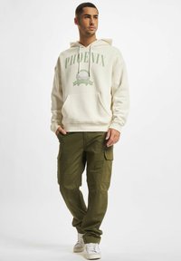 Felpa crema con grafica "PHOENIX", tasca a marsupio, abbinata a pantaloni cargo verde oliva con molteplici tasche e una vestibilità rilassata.