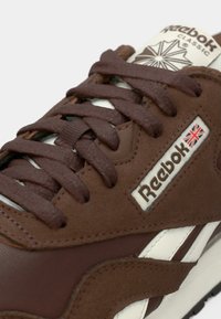 Zatvoreni plan smeđe Reebok Classic tenisice koja prikazuje teksturirane vezice, logo s Union Jack-om te detalje od antilop kože i kože.