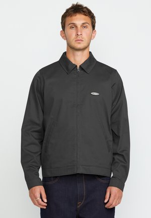 Chaqueta gris con cremallera y cuello puntiagudo, dos bolsillos frontales y un pequeño logo de la marca en el pecho. Tejido suave con textura lisa.