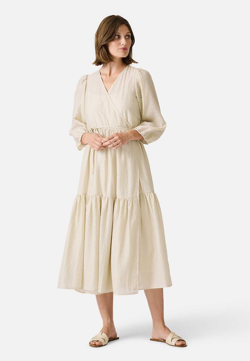 Krémové šaty do půli stehen s překládaným designem, výstřihem do V a dlouhými puff sleeves. Obsahují stupňovanou sukni a texturovanou látku s jemnými vzory.
