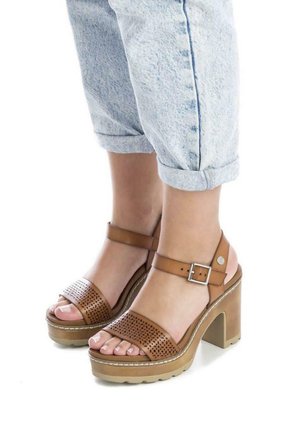Sandalias de plataforma de cuero marrón con parte superior perforada, correa ajustable en el tobillo y tacón grueso. Combinadas con pantalones de mezclilla claros de corte recto.