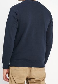 Laivastonsininen puuvillainen collegepaita, jossa pitkät hihat, pyöreä pääntie ja resorinen helma. Ominaisuuksiin kuuluu sileä koostumus ja ompelusaumat olkapäillä.