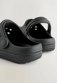 Melni plastmasas klogi ar atvērtām aizmugurēm, struktūrētām virsmām un vairākiem ventilācijas caurumiem. Ronde formas un plakana, pret slīdēšanu izturīga zole.