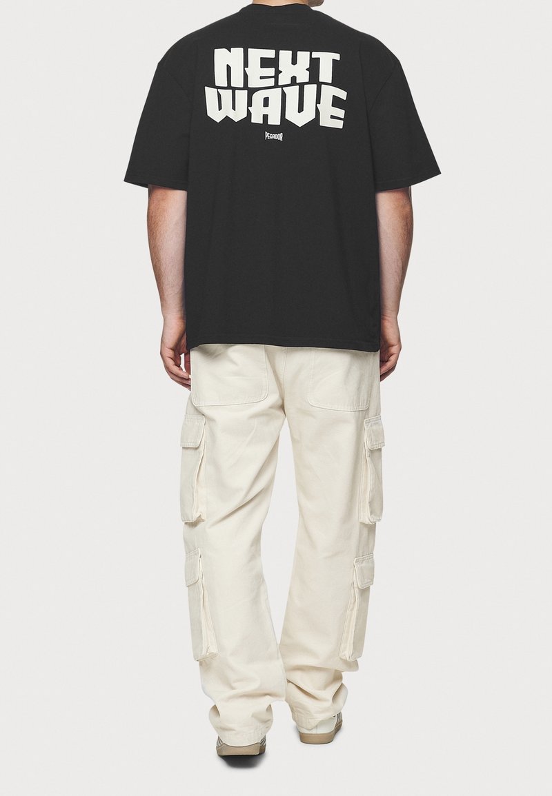 Czarny, oversize t-shirt z napisem "NEXT WAVE" w dużych, białych literach na plecach. Połączony z beżowymi spodniami cargo z wieloma kieszeniami.
