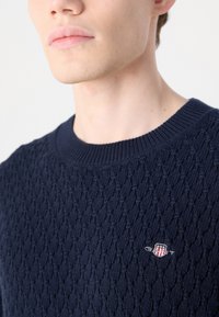 Meriharmaa neulepusero, jossa on teksturoitu timanttimäinen kuvio, ribbireunus kauluksessa ja pieni brodeerattu logo rinnassa.