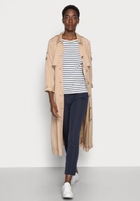 Beige trenchcoat med stora knappar, marinblå byxor och en vit-randig skjorta. Modellen bär vita slip-on skor; händerna avslappnade vid sidorna.