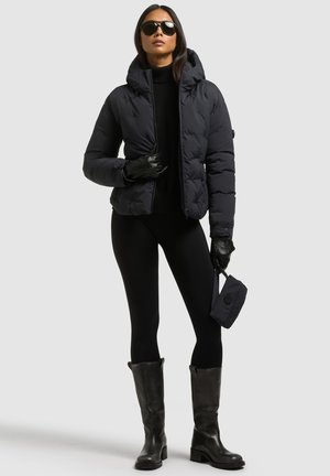 Un mannequin porte une doudoune noire, des lunettes de soleil, des gants, un legging et des bottes hautes jusqu’aux genoux, et tient un petit sac noir sur un fond gris uni.