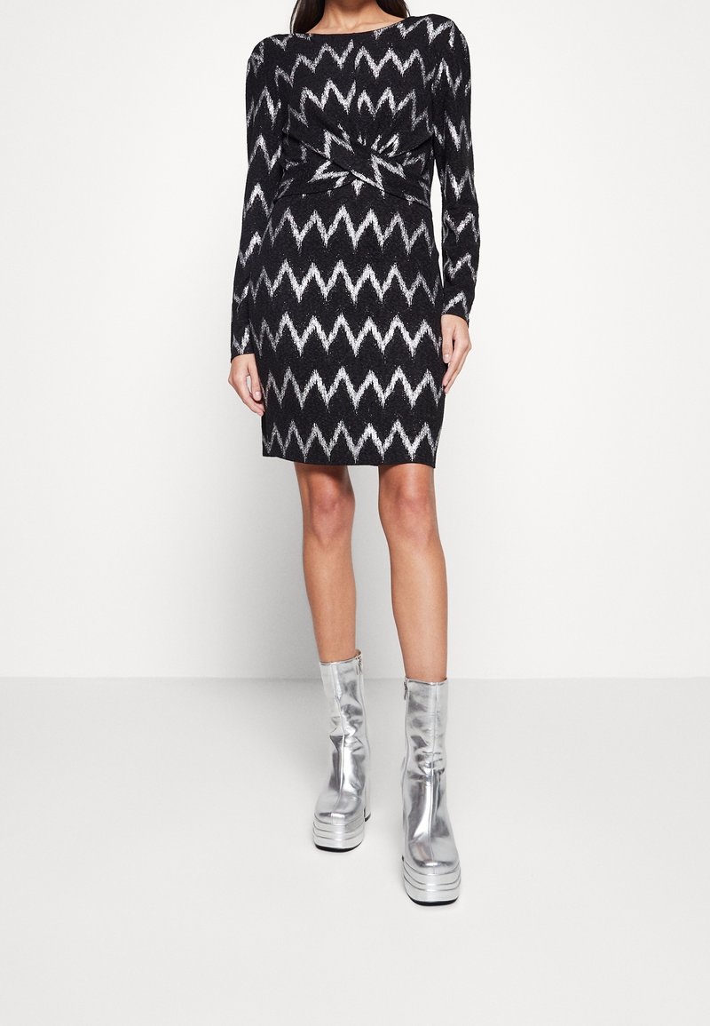 Robe noire et argent à motifs zigzag avec une taille tordue, manches longues, associée à des bottes plateforme argentées brillantes.