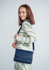 Tmavě modrá kožená taška přes rameno s klopnou, má prošívaný design a nastavitelný popruh, kterou nosí modelka v vzorovaném outfitu.