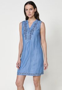 Robe en denim bleu clair avec un col mandarin, sans manches, ornée d'une broderie bleue complexe sur le devant et d'un ourlet en zigzag.