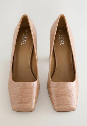 Kūno spalvos lakuotos odos aukštakulniai bateliai su stačiakampiu priekinių dalių padu, turintys krokodilo tekstūrą. Vidaus dalis yra lygi su įspaustu prekės ženklu ant vidpadžio.