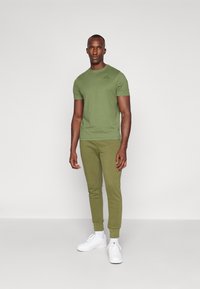 Πράσινη μπλούζα τύπου t-shirt και αντίστοιχες φόρμες από μαλακό ύφασμα; διαθέτει στρογγυλή λαιμόκοψη και μια διακριτική λεπτομέρεια με λογότυπο στο στήθος. Λευκά αθλητικά παπούτσια.