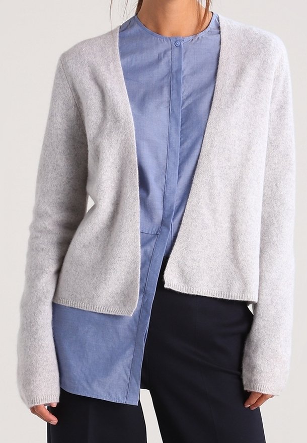 Femme portant un cardigan gris clair ouvert sur une chemise bleue boutonnée, associée à un pantalon noir.