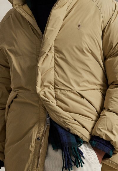 Bēšas krāsas pūderveida jaka ar rāvējslēdzēja aizdari un sānu kabatām. Uz krūtīm ir izšūts logotips, un jaka ir ar teksturētu izskatu, kurā redzamas šuvju detaļas.