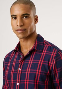 Camisa xadrez em vermelho e azul marinho, feita de algodão. Apresenta um colarinho abotoado e botões brancos, com um pequeno logótipo no peito.