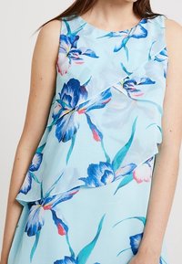 Robe bleu clair sans manches avec un motif floral en bleu et rose. Tissu doux avec volant superposé à la taille et encolure arrondie.