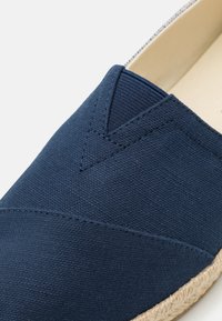 Mornarska platnena klizna cipela s teksturiranom gornjom stranom, V-oblika šavnog detalja i prirodnom jute užadnom potplatu. Unutra se nalazi lagana bež podstava.