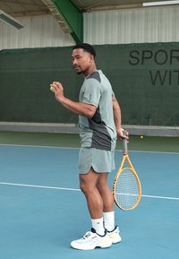 Conjunto desportivo em cinza com acentos escuros, apresentando uma parte de cima de manga curta e calções, combinado com sapatilhas brancas de ténis e uma raquete de ténis laranja.
