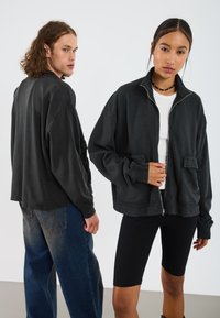 Crna zip-up dukserica sa opuštenim krojem, rebrastim manžetama i dva prednja džepa. Prikazana na dva modela koji nose ležerne outfite.