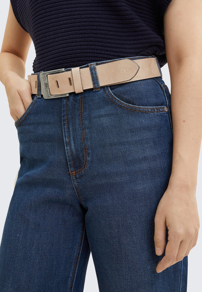 Brūns ādas jostas ar sudraba metāla sprādzi, uzvilkta virs tumši zila džinsu biksēm. Tuvplāns uz vidukļa, izceļot šuvju detaļas.