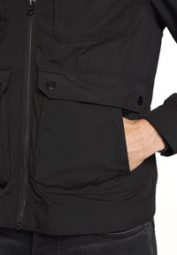 Main insérée dans la grande poche avant d'une veste noire zippée avec poches à rabat boutonnées et détails aux poignets.