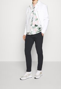Witte jas over een groen-roze shirt met patroon, gecombineerd met een donkere broek en lichte sneakers met een gaasstructuur en logo-accenten.