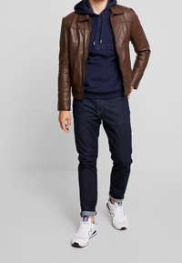 Brun læderjakke, marineblå hættejakke med logo, mørke jeans og hvide sneakers med marineblå detaljer. Casual outfit med lagdelte teksturer.