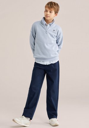 Zēns gaiši zilā ceturkšņa rāvējslēdzēja džemperī, svītrainā ar apkakli krekliņā, tumši zilās biksēs un baltos sporta apavos stāv ar rokām aiz muguras.