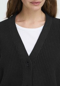 Cardigan noir en maille avec un col en V, présentant un motif texturé et une fermeture à un bouton, porté par-dessus un t-shirt blanc à côtes.
