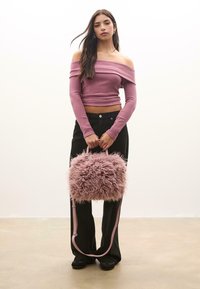 Lyserød fluffy håndtaske med tekstureret pelsfinish og kontrasterende lyserøde remme. Modellen bærer en tætsiddende off-shoulder ribbet top og sorte vide bukser.