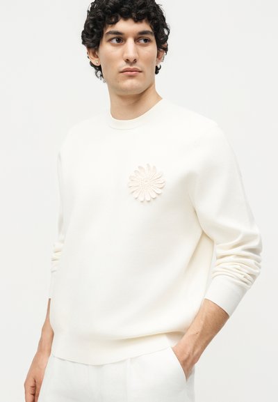 Άνδρας με σγουρά σκούρα μαλλιά που φοράει ολοκληρωμένο λευκό ντύσιμο με μεγάλη πλεκτή λεπτομέρεια λουλουδιού στο στήθος, κοιτάζοντας ελαφρώς προς το πλάι.